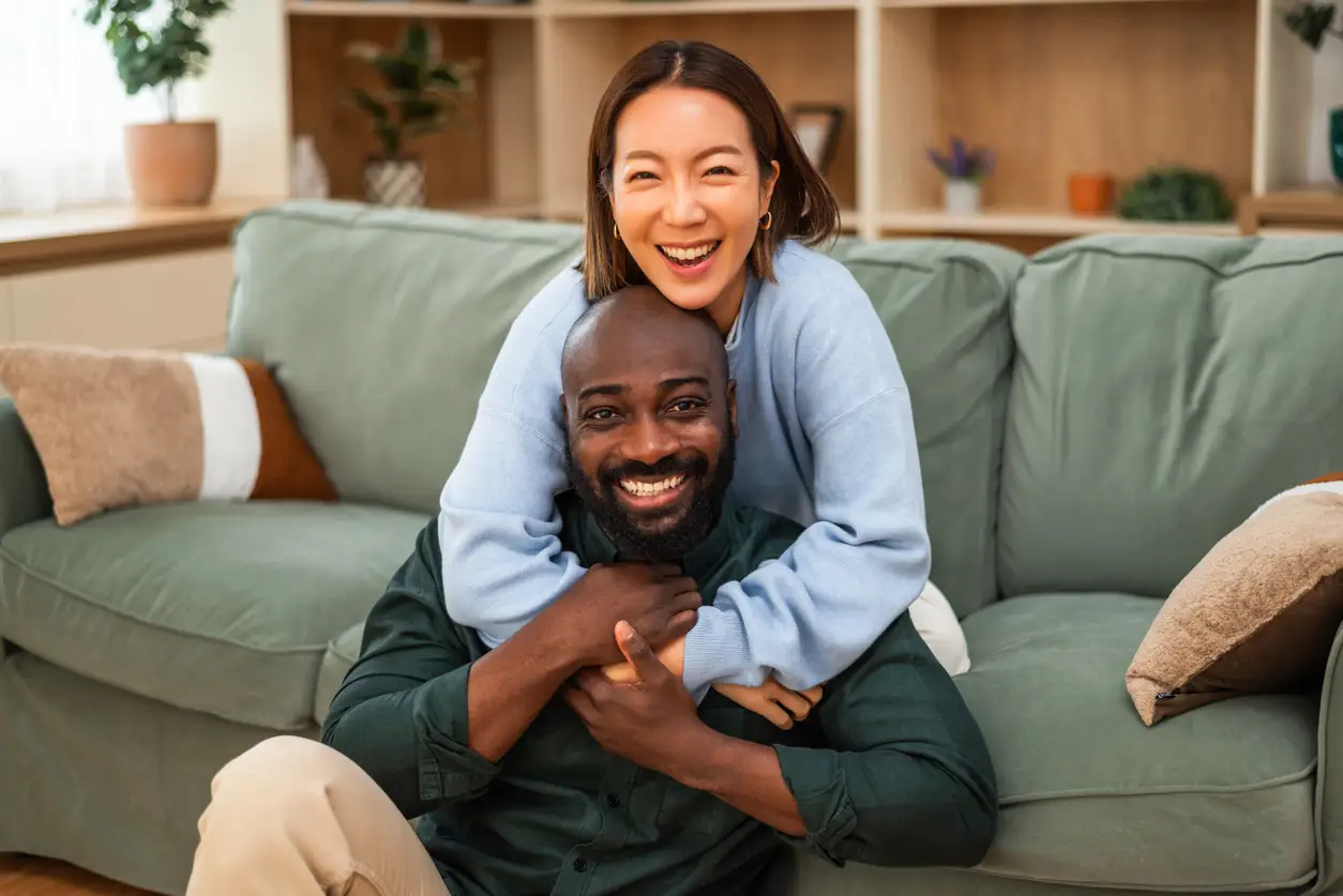 Cheerful interracial couple shares a joyful moment of laughter while relaxing at home. They are embracing and looking at camera.